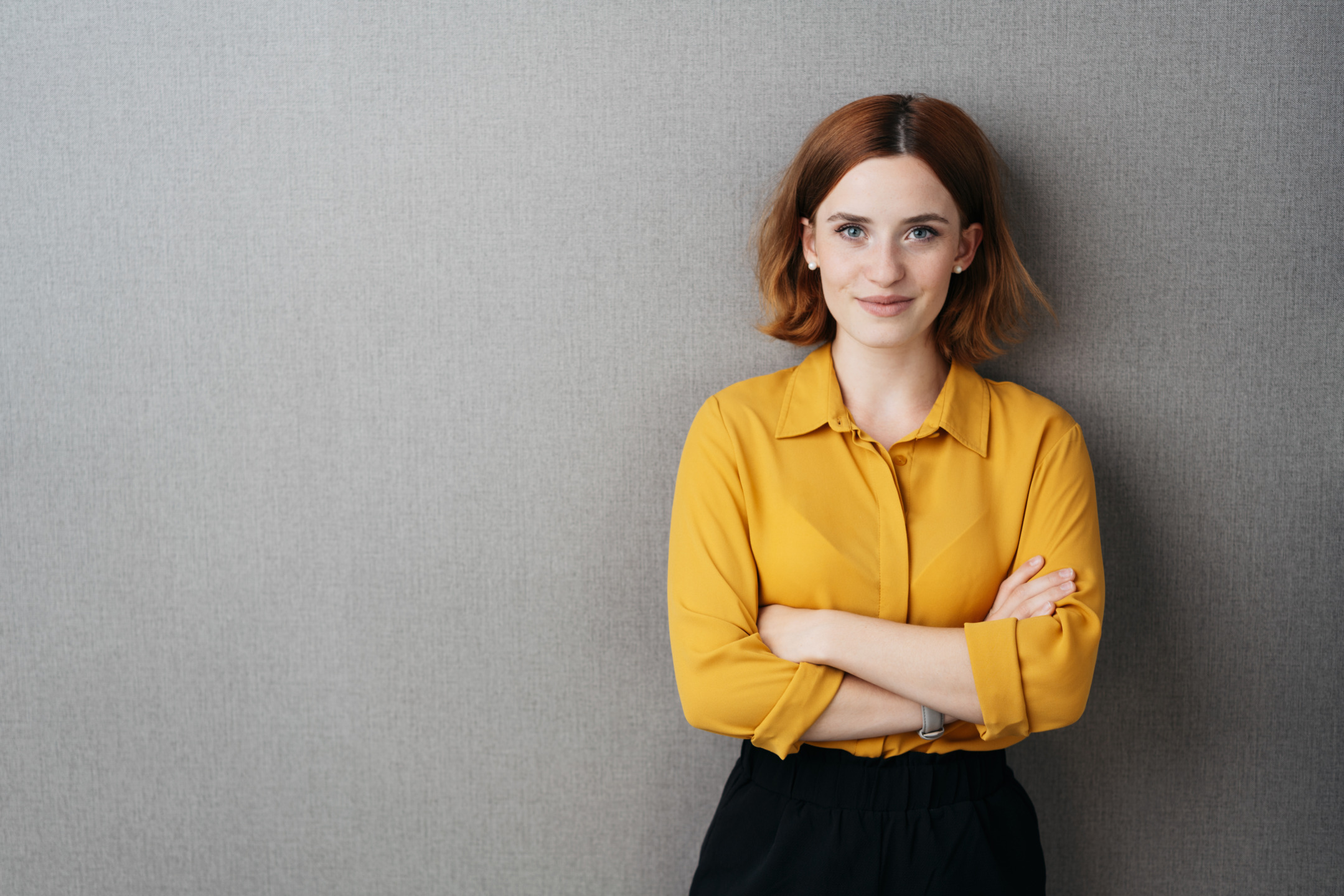 Friendly confident young woman standing with folded arms smiling at the camera over grey with copy space