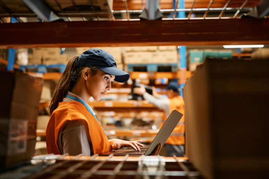 Female warehouse worker working on laptop at storage compartment.